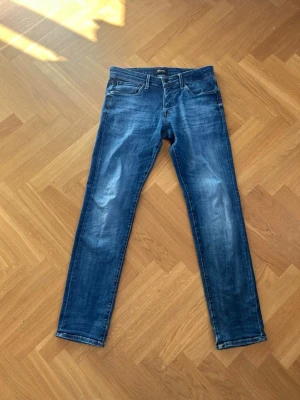 Blå slim fit jeans från Jack & Jones - Snygga blå slim fit jeans från Jack & Jones med klassisk design och diskret slitning på benen. Jeansen har en mörkblå tvätt och är tillverkade i bomull med stretch för extra komfort. Perfekt för dig som gillar en modern och stilren look. 