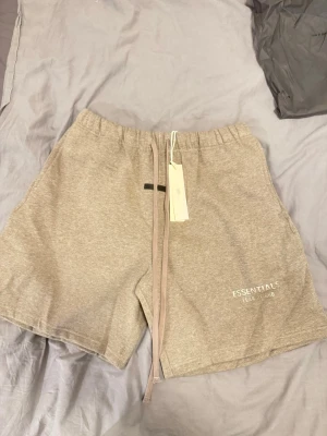 Beige Essentials shorts med snörning - Säljer ett par beige shorts från Fear of God Essentials med lång snörning i midjan och elastisk resår. Modellen är loose fit och har logotypen tryckt på ena benet. Perfekta för sommarens chill dagar.
