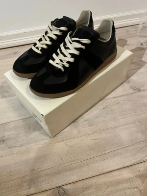 Maison Margiela Gats - Stilrena svarta sneakers med vita snören och bruna gummisulor. Skorna har en klassisk låg profil och är tillverkade i en mix av mocka och läder, vilket ger en snygg kontrast. Perfekta för dig som gillar minimalistisk och tidlös design. Skriv dm för frågor priset är inte fast!