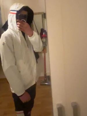 Moncler windbreaker vit - Säljer en vit jacka från Moncler med huva och dragkedja. Jackan har en snygg detalj med röd, vit och blå rand på huvan samt Moncler-logga på ärmen. Perfekt för dig som vill ha en stilren och exklusiv look.