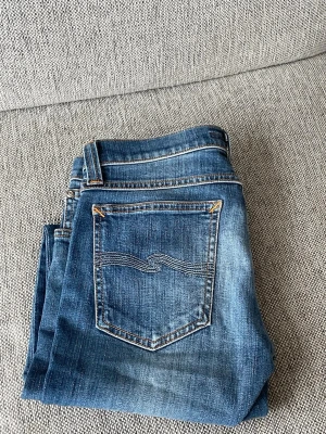 Blå slim fit nudie jeans - Snygga blå nudie slim fit jeans med slitna detaljer och ljusa partier på låren och knäna. Klassisk femficksmodell med dragkedja och knapp. Perfekta för en avslappnad och trendig look.   W28 L32