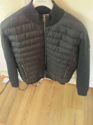 Svart pufferjacka från Moncler - Snygg svart pufferjacka från Moncler med quiltad front och släta ärmar. Jackan har hög krage, två dragkedjeförsedda fickor och diskret Moncler-logga på ärmen. Materialet är en mix av dun och syntet, perfekt för kalla dagar.