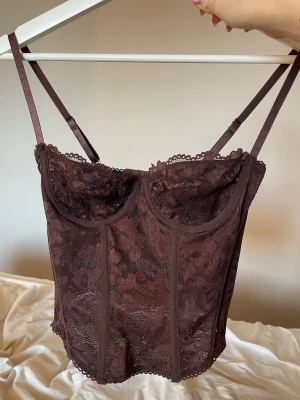 Brun spetskorsett med tunna axelband - Säljer en brun korsett med tunna, justerbara axelband och spetsdetalj över hela plagget. Korsetten har markerade sömmar, bygel under bysten och dekorativ kant upptill och nedtill. Stängs med hyskor i ryggen. 
