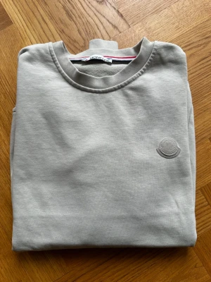 Beige sweatshirt från Moncler - Snygg beige sweatshirt från Moncler med rund halsringning och diskret Moncler-logga ton-i-ton på bröstet. Tillverkad i mjuk bomull för en skön och avslappnad look. Perfekt till jeans eller joggers för en clean stil. Lite stor i storleken och passar även M
