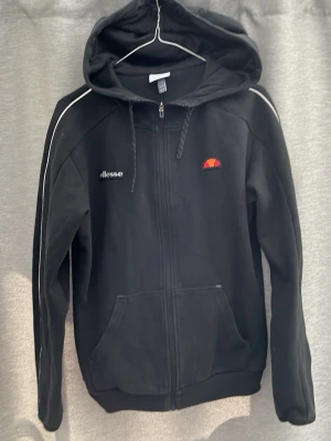 Svart hoodie från Ellesse med dragkedja - Snygg svart hoodie från Ellesse med dragkedja framtill och vita detaljer längs ärmarna. Klassisk huva med dragsko och två stora fickor framtill. Ellesse-logga broderad på bröstet. Perfekt för en avslappnad och sportig stil.