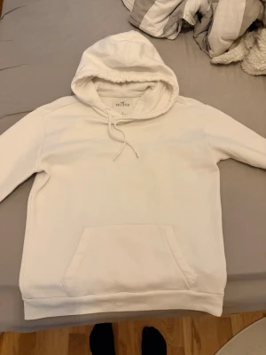 Vit hoodie från Hollister - Säljer en stilren vit hoodie från Hollister med klassisk känguruficka och dragsko i huvan. Hoodien är tillverkad i mjukt bomullsmaterial och har en relaxed fit som passar perfekt till chill dagar. Enkel och clean design utan tryck.