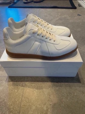 Vita sneakers från Maison Margiela Storlek 44 - Snygga vita sneakers från Maison Margiela med klassisk design och bruna gummisulor. Skorna har en låg profil, vita läderpaneler och matchande vita skosnören. Perfekt för dig som gillar stilrena och trendiga sneakers. Storlek 44 men passar även 43