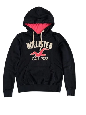 Mörkblå Hollister hoodie med brodyr - Svart hoodie från Hollister med broderad logga och text 'CALI. 1922' på bröstet. Insidan av huvan är rosa och dragsnörena är vita. Klassisk känguruficka framtill och ribbade muddar. Perfekt för en avslappnad och cool stil.