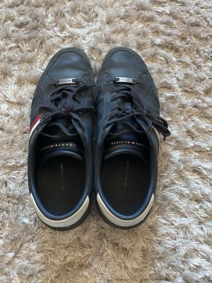 Svarta Tommy Hilfiger sneakers - Snygga svarta sneakers från Tommy Hilfiger med klassiska röda och vita detaljer på sidan. Skorna har låga skaft, svarta snören och en vit hälkappa. Materialet är en mix av läder och textil, perfekt för en clean och stilren look.