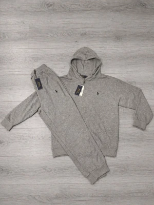 Polo Ralph Lauren Set 🩶 - Snygg grå hoodie från Polo Ralph Lauren med klassisk huva och diskret logga broderad på bröstet. Tillverkad i mjukt material som känns skönt mot huden. Perfekt för en avslappnad och stilren look. Matchande byxor ingår ej. Storlek XS och L i barnstorlek 