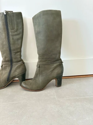 Olivgröna höga mockastövlar med klack - Snygga höga stövlar i olivgrönt skinn med rundad tå och bred klack. Stövlarna har dragkedja på insidan och en stilren design som passar till många outfits. Perfekta för dig som vill ha både höjd och komfort. Supersköna! Står storlek 37 men passar mig med 37,5-38!