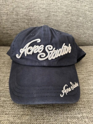 Acne studios keps - Snygg mörkblå keps från Acne Studios med broderad logga i vitt framtill och på skärmen. Klassisk böjd skärm och justerbar passform bak. Tillverkad i bomull för en bekväm känsla och stilren look.