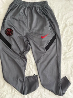 Gråa PSG träningsbyxor från Nike - Snygga gråa träningsbyxor från Nike med PSG-logga och röd Nike Swoosh på framsidan. Byxorna har svarta detaljer, elastisk midja och dragkedja vid benslut. Perfekta för träning eller chill. Materialet är mjukt och stretchigt, troligen polyester.