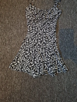 Svart blommig kortklänning - Söt svart kortklänning med vita blommor och smala axelband. Klänningen har en figurnära passform upptill och faller ut i en lätt utställd kjol. Perfekt för varma dagar och enkel att styla med sneakers eller sandaler. Och har inbyggda shorts. 