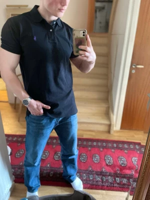 Svart piké från Polo Ralph Lauren - Svart piké från Polo Ralph Lauren i 100% bomull. Klassisk design med krage, knappar framtill och korta ärmar. Liten broderad logga på bröstet ger en stilren touch.