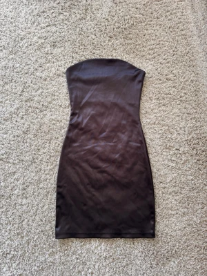 Brun axelbandslös kort klänning - Snygg brun axelbandslös klänning i kort modell med figurnära passform. Klänningen har en enkel och stilren design, perfekt för dig som gillar minimalistiskt mode. Tillverkad i ett mjukt och stretchigt material som sitter bekvämt.