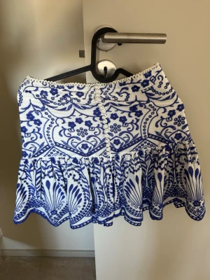 Vit och blå mönstrad kjol från Zara - Supersöt kort kjol från Zara i vitt med blått mönster och dekorativa knappar framtill. Kjolen har volang nertill och ett unikt, nästan bohemiskt mönster. Perfekt för dig som gillar att sticka ut med detaljer och färg!