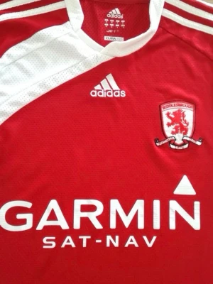 Middlesbrough Adidas matchtröja röd - Röd och vit matchtröja från Adidas för Middlesbrough FC med klubbmärke på bröstet och Garmin som sponsortryck. Tröjan har vita detaljer på axlar och sidor, samt klassiska Adidas-ränder. Tillverkad i lätt och ventilerande funktionsmaterial.