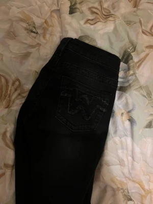 Svarta jeans från Wrangler - Säljer ett par svarta jeans från gina tricot med klassisk femficksmodell och tydlig W-detalj på bakfickan. Jeansen har vida ben och silverfärgade knappar. Perfekta för en cool och avslappnad stil. Helt oanvända eftersom de tyvärr är för korta. 