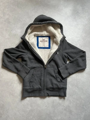 Hollister Hoodie - Mysig mörkgrå hoodie från Hollister med dragkedja och vit fluffig teddyfoder på insidan. Stor logga på bröstet och två fickor framtill. Perfekt för dig som vill ha en varm och stilren hoodie med huva.