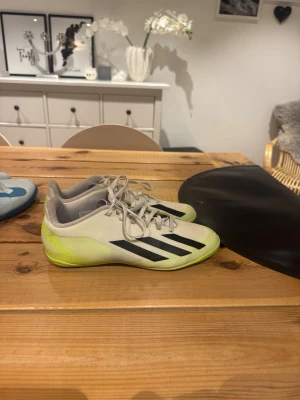 Adidas vita och gula fotbollsskor - Säljer ett par vita och ljusgula fotbollsskor från Adidas med svarta ränder på sidan. Skorna har snörning och platt sula med mönster för bra grepp. Perfekta för dig som vill ha snygga och funktionella skor på planen.