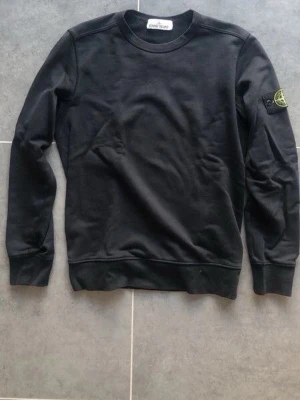 Svart långärmad tröja från Stone Island - Svart långärmad tröja från Stone Island med rund halsringning och klassisk logotyp-patch på ena ärmen. Tröjan har en enkel och stilren design i mjukt material, perfekt för en avslappnad look.