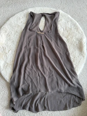 Gråbrun ärmlös linnetopp med metall-detalj - Säljer en gråbrun ärmlös linnetopp från Fashion med snygg metall-detalj vid halsen och öppning i ryggen. Toppen har en luftig passform och är längre baktill, perfekt för att skapa en cool och avslappnad look.