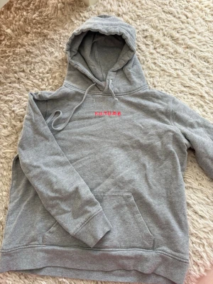 from future hoodie - Snygg grå hoodie med huva och dragsko, samt magficka framtill. På bröstet finns broderad texten F.U.T.U.R.E i rosa. Perfekt för en chill och avslappnad stil. Mjuk och skön bomullsblandning.