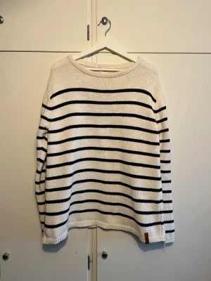 Randig stickad tröja Knowledge Cotton - Säljer en vit stickad tröja med svarta horisontella ränder från Knowledge Cotton Apparel. Tröjan är gjord av mjuk ekologisk bomull och har rund hals samt ribbade muddar. Perfekt för dig som gillar klassisk och stilren design.