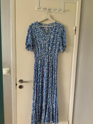 Blommig långklänning  - Långklänning i blått med vitt blommönster, v-ringad och med korta puffärmar. Klänningen har en smickrande passform och volangkant nertill. Perfekt för sommarens alla tillfällen.