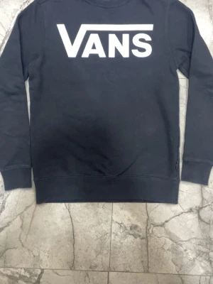 Svart sweatshirt från Vans - Svart långärmad sweatshirt från Vans med stor vit logga framtill. Klassisk rund halsringning och ribbade muddar vid ärmslut och nederkant. Perfekt för en avslappnad streetwear-look.