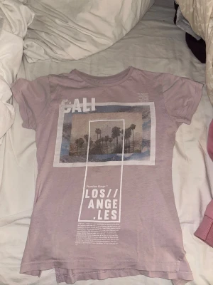 Ljuslila t-shirt med LA-tryck - tröjan har inga hål eller fläckar, men det är två små snitt längst ner på sidorna av tröjan(den är köpt så). skönt material. storleken är 140/ 9-10 years men jag har storlek 170 och är 13 år och den passade bra.