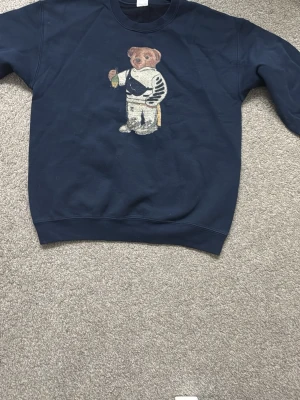 Mörkblå sweatshirt med björnmotiv - Cool mörkblå sweatshirt med ett broderat björnmotiv på bröstet. Tröjan har rund halsringning och ribbade muddar vid ärmslut och nederkant. Perfekt för dig som gillar unika detaljer och streetwear-vibe.