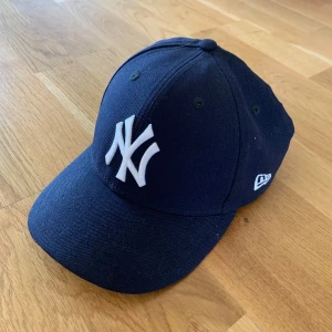 Mörkblå New Era Yankees keps - Snygg mörkblå keps från New Era med klassiska New York Yankees-loggan broderad framtill i vitt. Kepsen har böjd skärm, ventilationshål och broderad New Era-logga på sidan. Perfekt accessoar för en sportig och trendig stil.