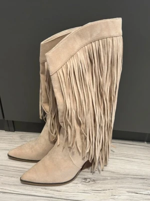 Beiga cowboy boots med fransar - Snygga beiga cowboy boots med långa fransar längs skaftet och spetsig tå. Bootsens material ser ut att vara mocka och de har en klassisk westernstil med låg klack. Perfekta för dig som vill sticka ut med en trendig look. Storlek 40