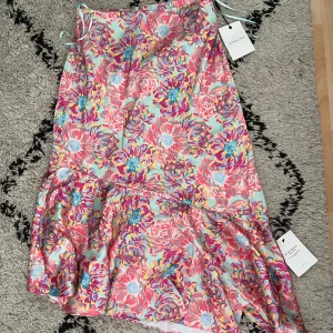 Blommig kjol från By Malina - Säljer en färgglad blommig kjol från By Malina i storlek XS. Klänningen har volangdetalj nertill och är i ett glansigt material med rosa, blå och gula blommor på ljus bakgrund. Perfekt för sommarens tillfällen!