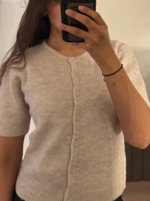 Beige kofta med kort ärm - Mysig beige kofta med korta ärmar och rund halsringning. Koftan har knäppning framtill med matchande knappar och ribbade muddar vid ärmslut och nederkant. Perfekt för lager-på-lager och enkel att styla till olika outfits.