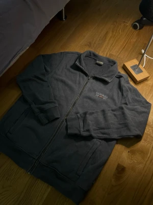 Mörkgrå ziptröja från Napapijri - Mörkgrå stickad ziptröja från Napapijri med broderad logga och årtalet 1987 på bröstet. Tröjan har hög krage, hel dragkedja och två fickor framtill. Perfekt för lager-på-lager och chill vardagsstil.