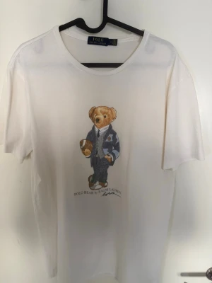 Vit Polo Bear t-shirt från Ralph Lauren - Vit t-shirt från Polo Ralph Lauren med ikoniskt Polo Bear-tryck framtill. Klassisk rund hals och korta ärmar. Tillverkad i mjuk bomull som känns skön mot huden. Perfekt för en avslappnad och stilren look.