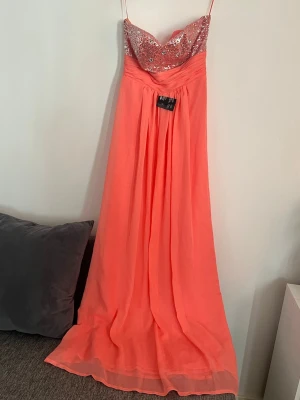 Balklänning  - Supersnygg långklänning i korallrosa med axelbandslös och glittrig paljettöverdel. Klänningen har ett svepande fall och markerad midja, perfekt för bal eller fest. Överdelen är dekorerad med silverfärgade paljetter som ger extra wow-känsla.
