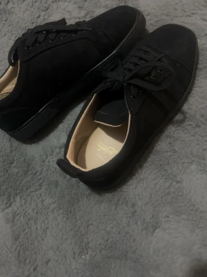 Svarta sneakers från Christian Louboutin - Stilrena svarta sneakers i mocka från Christian Louboutin med klassisk låg profil och svarta skosnören. Skorna har en ikonisk röd sula och diskreta detaljer som ger en lyxig känsla. Perfekta för dig som vill ha en clean och exklusiv look.