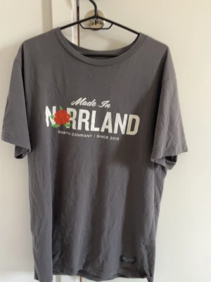 Grå Norrland T-shirt  - Snygg grå t-shirt från SQRTN Company med trycket 'Made in Norrland' och en röd-blommig detalj framtill. Klassisk rund hals och korta ärmar. Tillverkad i mjuk bomull, perfekt för en avslappnad stil.