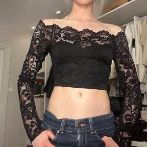 Svart croppad spetsblus offshoulder - Snygg svart croppad blus med offshoulder-design och långa ärmar i genombruten spets. Blusen har ett romantiskt spetsmönster och sitter tight över kroppen. Perfekt för dig som vill ha en trendig och elegant topp med unik detalj.