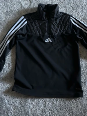 Svart träningsjacka från Adidas - Svart långärmad träningsjacka från Adidas med quiltade detaljer på axlarna och bröstet. Jackan har halv dragkedja framtill och klassiska vita Adidas-ränder längs ärmarna. Materialet är syntetiskt och passar perfekt för sport och träning.