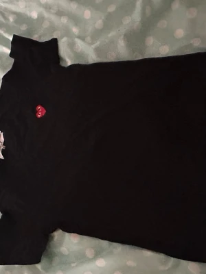 Svart t-shirt med rött hjärta - Svart t-shirt från Comme des Garçons Play med ett ikoniskt rött hjärta broderat på bröstet. Klassisk passform och korta ärmar, perfekt för en cool och enkel look. Materialet är mjuk bomull som känns skönt mot huden.