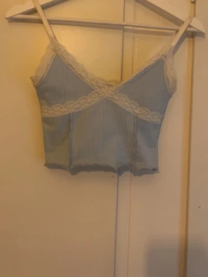 Ljusblå ribbad linnetopp med spets - Söt ljusblå ribbad linnetopp med tunna axelband och dekorativ vit spets längs urringningen och kanten. Toppen har en croppad passform och subtila vertikala ränder för extra detalj. Perfekt att styla med jeans eller kjol. Toppen har även justerbara axelband. Men på bild 4 kan du se att det är några trådar men inget som man tänker på.