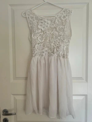 Vit spetsklänning med öppen rygg - Söt vit kortklänning med spets upptill och flowy kjol i chiffongliknande material. Klänningen är ärmlös och har en snygg öppen rygg med knappdetalj. Perfekt för sommarens alla tillfällen!