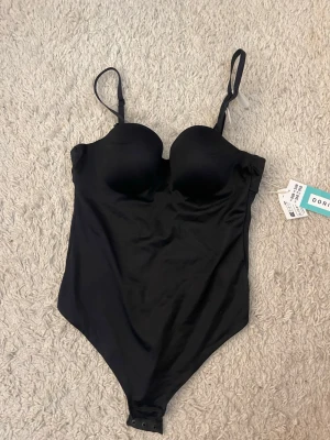 Svart body från Dorina med bygel - Snygg svart body från Dorina med vadderade kupor och bygel. Justerbara axelband och knäppning i grenen. Endast testad, storlek 80b