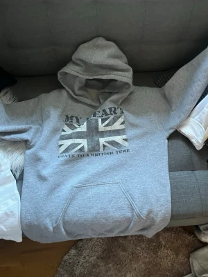 Grå hoodie med brittisk flagga Gildan - Snygg grå hoodie från Gildan med stor brittisk flagga och texten 'MY HEART BEATS TO A BRITISH TUNE' på bröstet. Klassisk känguruficka framtill och stor huva. Perfekt för dig som gillar streetwear och vill ha en chill look.