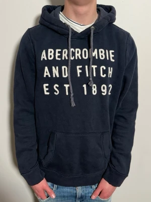 Mörkblå hoodie från Abercrombie & Fitch - Mörkblå vintage hoodie med ett snyggt tryck från Märket Abercrombie & Fitch. Storlek:S!! Skick8/10. Passar perfekt nu till varmare väder. Modellen är 176cm och väger 65kg. 